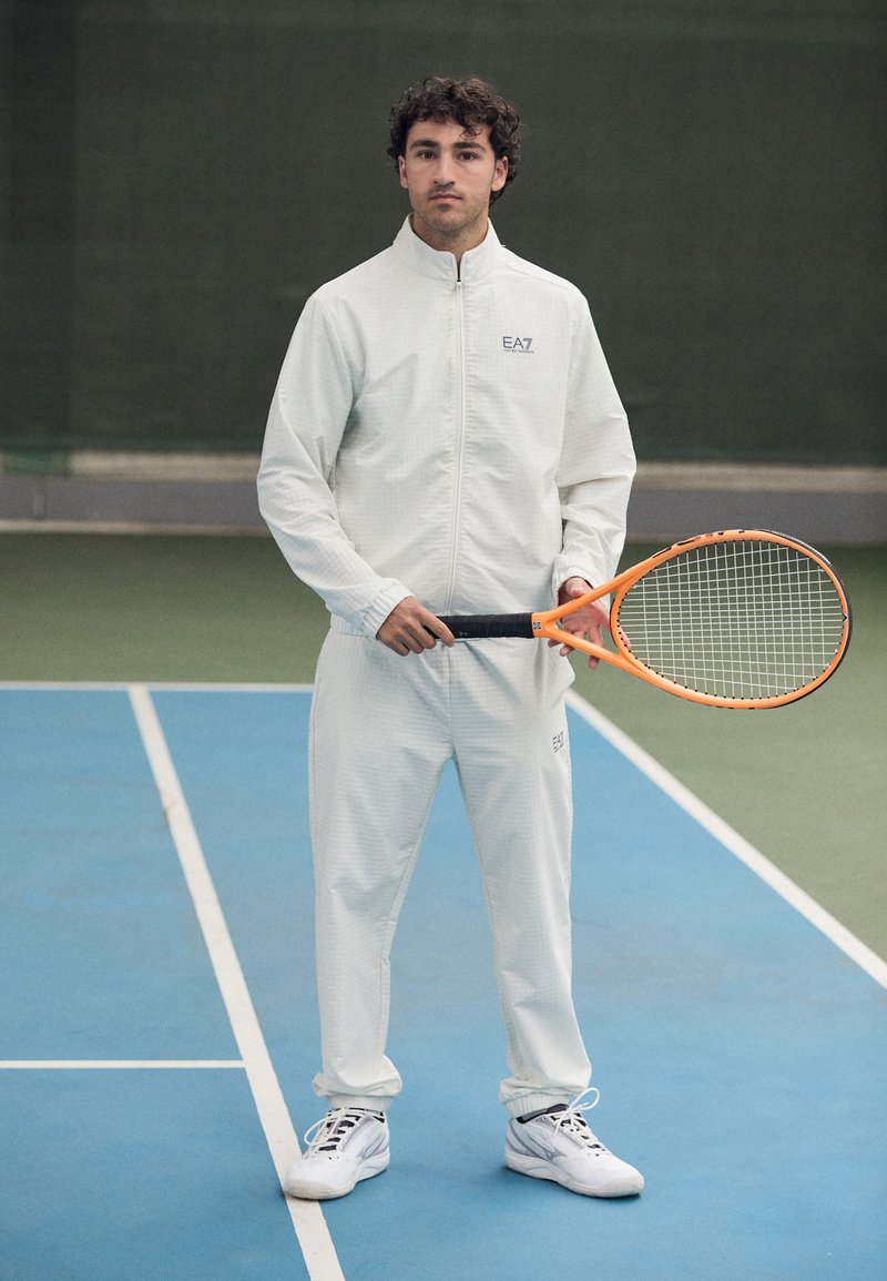 Fato de treino branco com padrão texturizado, apresentando fecho de correr e logótipo EA7. A segurar uma raqueta de ténis laranja num court azul.