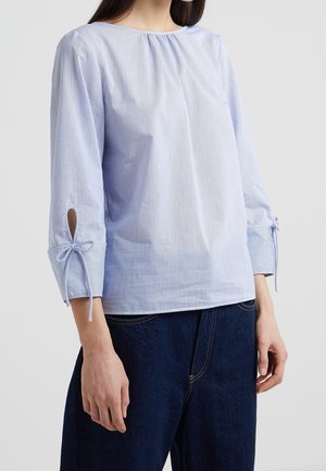 Vrouw draagt een lichtblauw gestreept blouse met driekwartmouwen, gestrikt bij de manchetten, gecombineerd met donkerblauwe spijkerbroek.