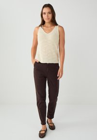 Beige mouwloos gebreid topje, met V-hals, gecombineerd met donkere bruin taps toelopende broeken. Model draagt zwarte perforated ballerina's. Eenvoudige en casual outfit.