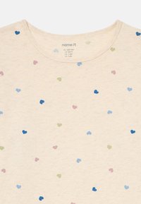 Ανοιχτό μπεζ T-shirt με στρογγυλή λαιμόκοψη, που διαθέτει ένα διασκορπισμένο σχέδιο από μικρές καρδιές σε μπλε, πράσινο και ροζ. Κατασκευασμένο από μαλακό υφασμάτινο υλικό.