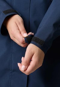 Giacca blu navy con primo piano delle mani che fissano il cinturino in Velcro al polsino. Il tessuto appare liscio e resistente.