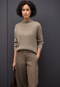 Beige strukturierter Pullover mit hohem Kragen und lockerer Passform, kombiniert mit braunen gewobenen Hosen. Das Model hält eine braune Tasche, vor einem dunklen Hintergrund.