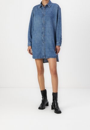 Personne portant une robe chemise en denim bleu avec un texte blanc répété et des bottines noires à talons, debout devant un fond blanc.