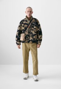 Fleece pullover with a black and beige floral pattern, paired with tan pants. Accessories include a small beige crossbody bag and white sneakers.