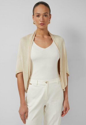 Vrouw draagt een crèmekleurige korte mouwen vest over een witte mouwloze top en witte broek met hoge taille, staande voor een effen grijze achtergrond.