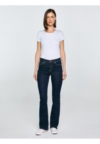 Žena stoji okrenuta prema naprijed, nosi jednobojnu bijelu majicu, tamnoplave traperice i bijele tenisice na svijetlosivoj pozadini.