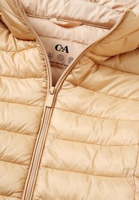 Béžová prošívaná bunda s částečně rozepnutým zipem, která ukazuje štítek značky C&A a ikony pro recyklované materiály, lehkost a možnost praní v pračce.