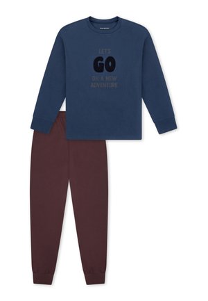 Lächelnder Junge mit blondem Haar steht barfuß mit den Händen hinter dem Kopf, trägt ein blaues Langarmshirt und braune Hosen vor weißem Hintergrund.