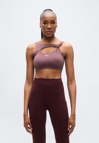 Reggiseno sportivo Nike color malva con ritaglio asimmetrico e spallina regolabile; abbinato a leggings bordeaux a vita alta con tasche laterali.