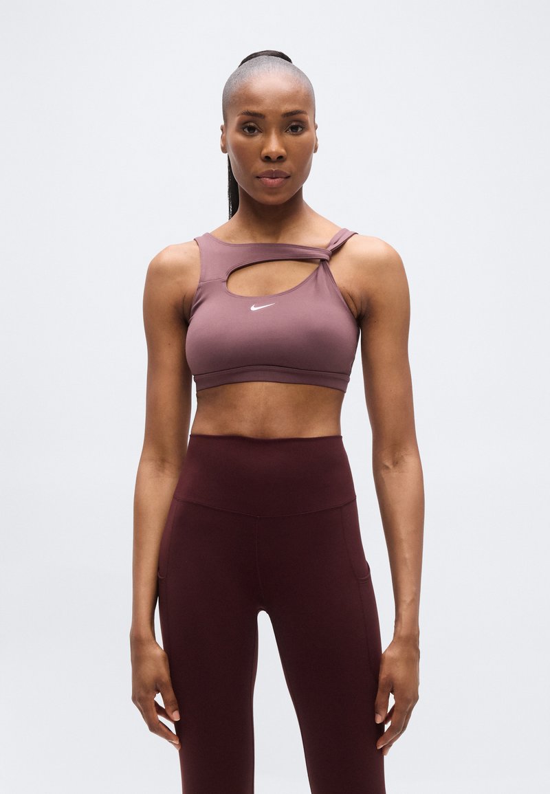 Reggiseno sportivo Nike color malva con ritaglio asimmetrico e spallina regolabile; abbinato a leggings bordeaux a vita alta con tasche laterali.