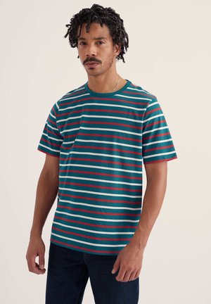 Göndör hajú férfi, akinek türkizkék, piros és fehér csíkos rövid ujjú inget és sötét nadrágot visel, egyszínű világos háttér előtt áll.