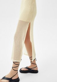 Pletené béžové šaty s rozparky po stranách, doplněné černými sandály na vysokém podpatku s silnou podrážkou a křížovými řemínky kolem kotníků.