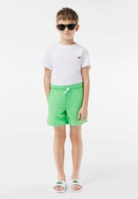 Kind in een wit T-shirt met korte mouwen met een klein logo, groene shorts en witte sandalen met groene accenten, gecombineerd met donkere zonnebril.