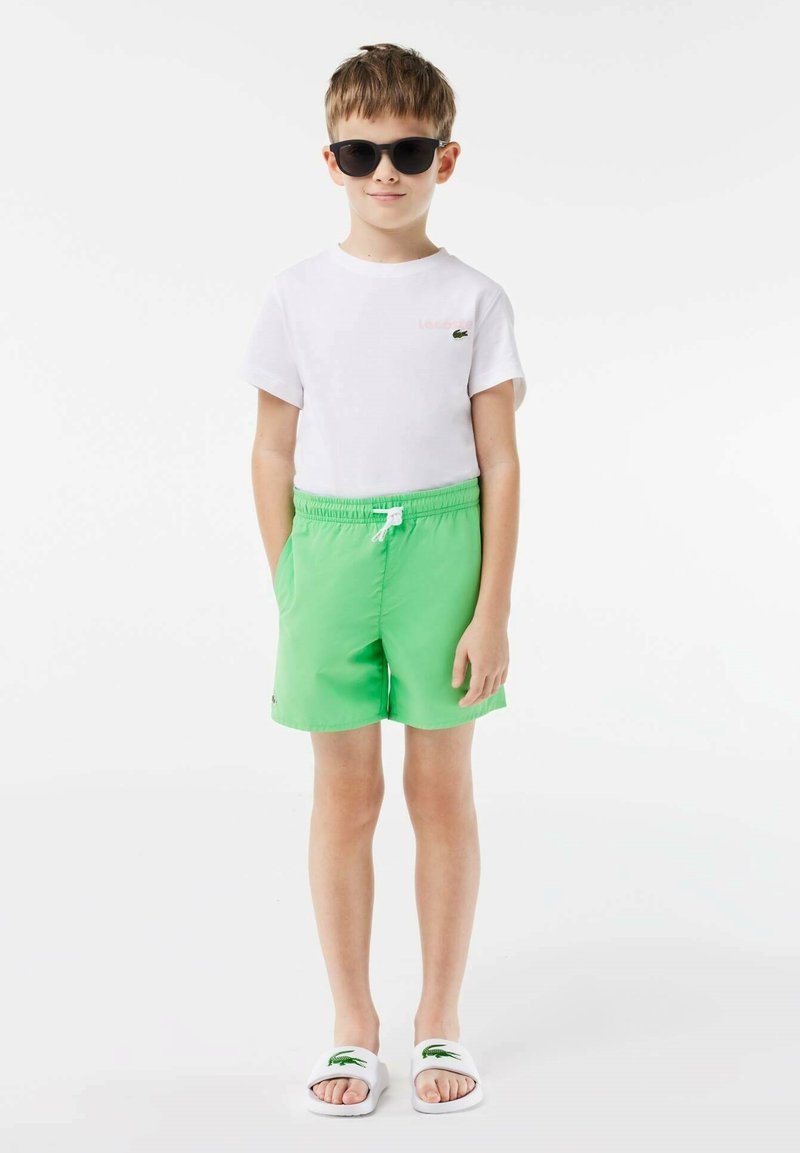 Kind in een wit T-shirt met korte mouwen met een klein logo, groene shorts en witte sandalen met groene accenten, gecombineerd met donkere zonnebril.