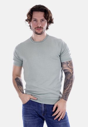 Suéter de punto de manga corta verde claro con cuello redondo, diseño ajustado y dobladillo acanalado. El modelo lleva puesta jeans azul oscuro y muestra tatuajes.