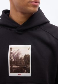 Κοντινό πλ άνο ενός ατόμου που φοράει μαύρο φούτερ με κουκούλα, με σχέδιο παλιού στυλ βουνού και δάσους και το κείμενο "HUGO" σε κόκκινο στο στήθος.