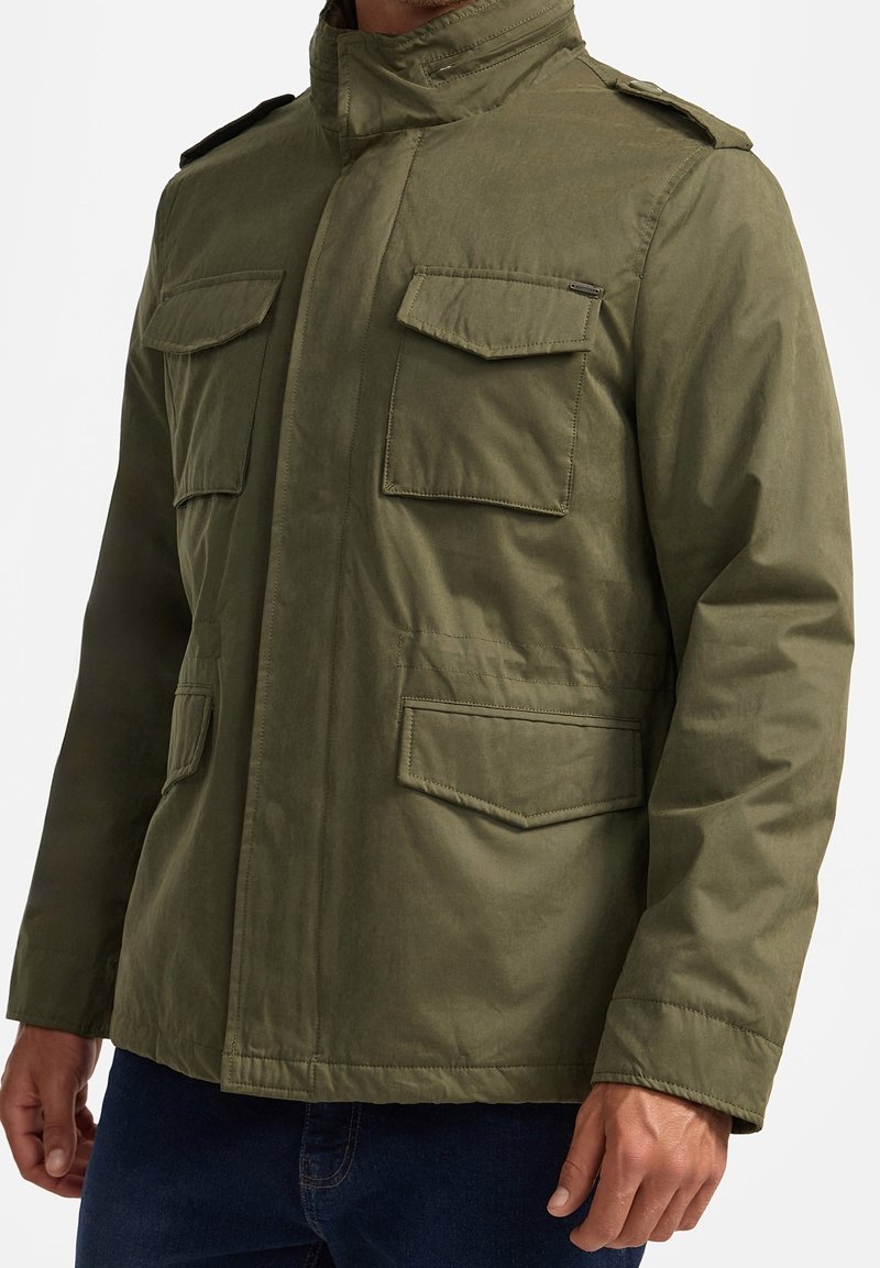 Homme portant une veste style militaire vert olive avec quatre poches à rabat et des épaulettes, associée à un jean bleu foncé.