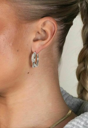 Silver hoopsörhängen med sammanflätad design, med en strukturerad yta och släta kanter. Den glänsande ytan kompletterar bärarens hudton.