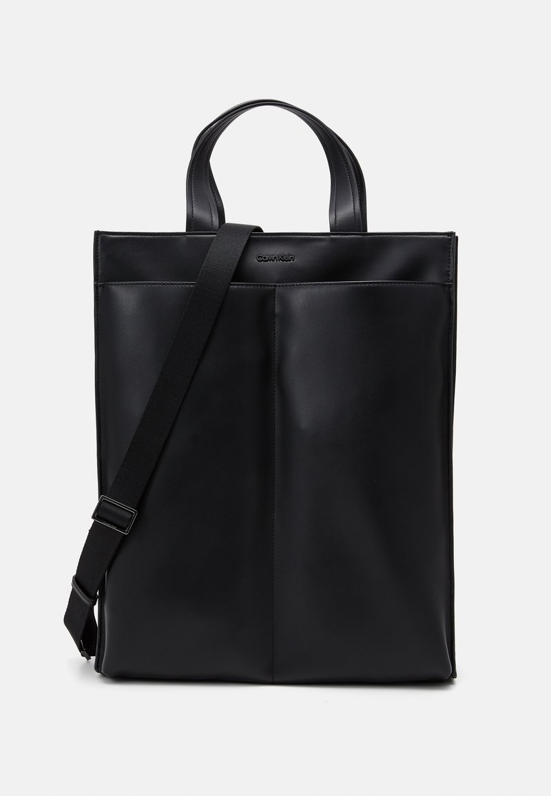Sac cabas noir Calvin Klein avec deux anses, une bandoulière amovible et une grande poche avant, sur un fond uni.