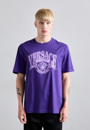Ung man med blont hår iklädd en lila Versace T-shirt med ett Medusa-huvud och lagerkransdesign, stående mot en enfärgad vägg.