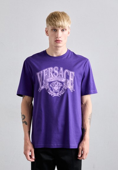 Młody mężczyzna o blond włosach, ubrany w fioletową koszulkę Versace z motywem głowy Meduzy i wieńcem laurowym, stojący przy gładkiej ścianie.
