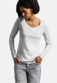 Vit långärmad topp med rundad halsringning, gjord av mjukt tyg. Kombinerad med ljusgrå jeans, händerna placerade i fickorna.