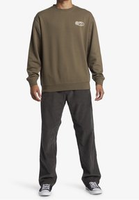 Olivově zelená mikina s kulatým výstřihem a bílým logem, kombinovaná s tmavě šedými kalhotami se širokými nohavicemi a černými teniskami s bílými detaily.