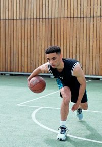 Basketball player in black sleeveless shirt and teal shorts dribbling a ball on a court with green flooring and wooden backdrop.