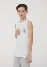 Maglietta bianca senza maniche con scollatura rotonda e grafica nera "LA" sul petto sinistro, abbinata a pantaloni sportivi grigi. Texture del tessuto liscia.