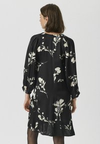 Robe noire avec motif floral blanc, manches longues et coupe décontractée. Le dos présente un détail de bouton au niveau du col et un léger brillant.