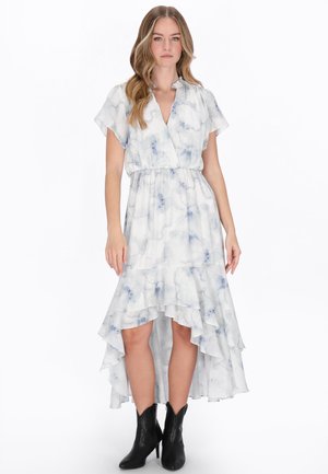 Donna che indossa un vestito bianco e grigio tie-dye a taglio asimmetrico con maniche corte e stivali neri alla caviglia, in piedi su sfondo bianco.