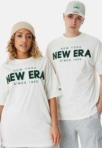 Zwei Personen tragen übergroße weiße T-Shirts mit grünem "NEW ERA"-Text und einem Logo. Die Frau trägt eine beigefarbene Mütze, der Mann eine graue Cap.