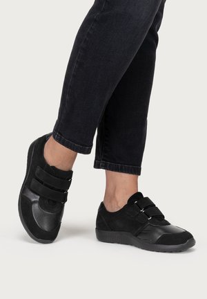 Person trägt schwarze Sneakers mit doppelten Klettverschlüssen und schwarze kurze Jeans und steht auf hellem Hintergrund.