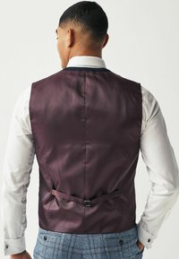 Burgundská saténová vesta so slim fit strihom, s prackou na úpravu vzadu, v kombinácii s bielou košeľou a károvanými nohavicami.