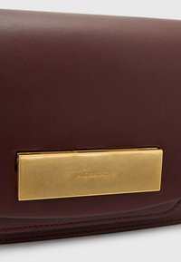 Mørkerød læder clutch-pung med en gylden rektangulær spænde med prægede brandnavn. Glat tekstur med et rent, minimalistisk design.
