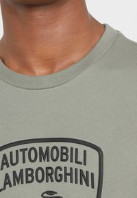Camiseta de cuello redondo color verde oliva, fabricada en algodón, con un gran logo negro en relieve de "AUTOMOBILI LAMBORGHINI" en el frente. Textura suave.