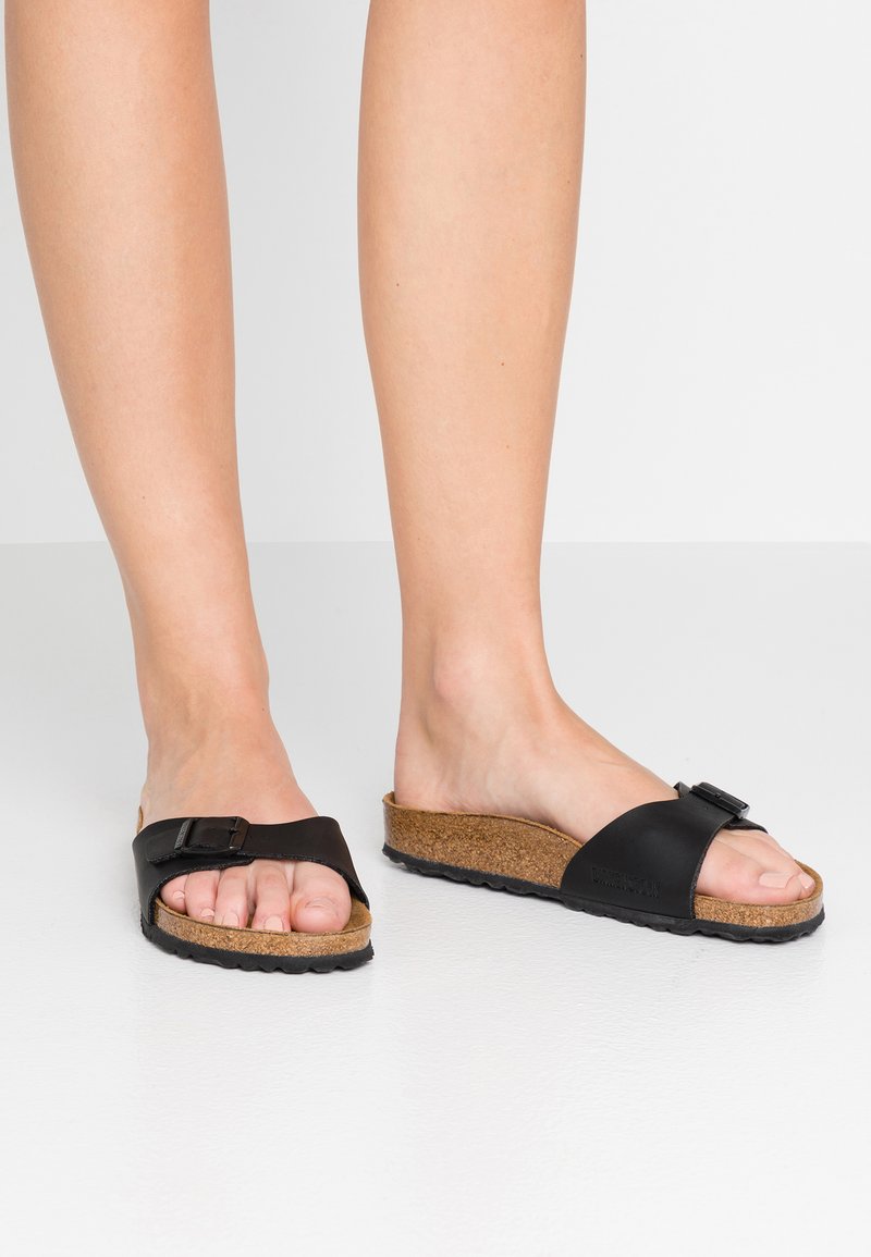 Sorte slidesandaler har en korkfodersål, flad sål og en bred rem med en spænde for et sikkert fit. Tekstureret ydersål for godt greb.