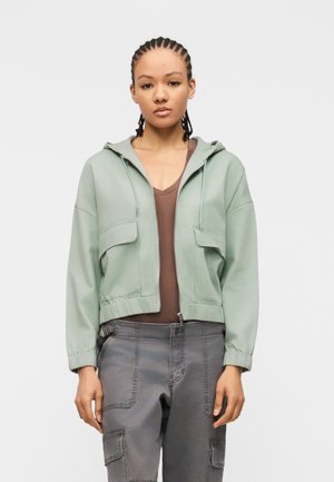 Mladá osoba s vrkočmi, oblečená v svetlozelenom kapucňovom kabáte cez hnedý top a sivé pracovné nohavice, stojaca pred jednoduchým pozadím.