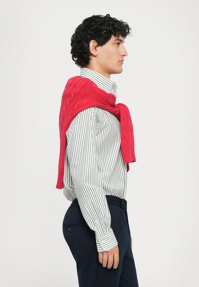 Pulover tricotat roșu lăsat peste o cămașă cu dungi albastre și albe, asociat cu pantaloni bleumarin. Caracteristicile includ un guler clasic și detalii cu nasturi.