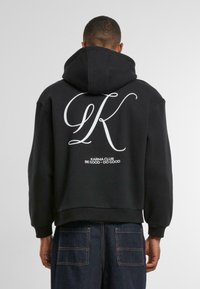 Zwarte hoodie met een trekkoordkap, met een grote witte "LK" grafiek en de tekst "KARMA CLUB BE GOOD - DO GOOD" op de achterkant.