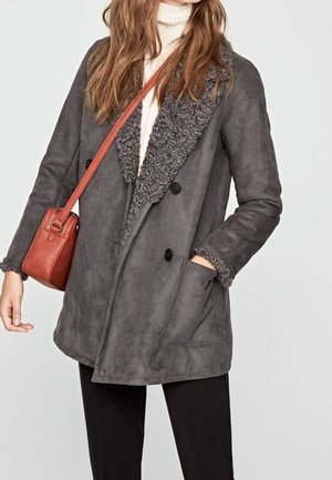 Mujer con un abrigo gris de borrego sobre un suéter de cuello alto blanco, pantalones negros y llevando un pequeño bolso cruzado de cuero marrón.