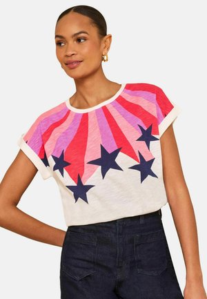 Frau mit goldenen Creolen, die ein weißes T-Shirt mit roten und pinkfarbenen Strahlen und großen dunkelblauen Sternen trägt, kombiniert mit einer dunklen, hoch geschnittenen Hose.