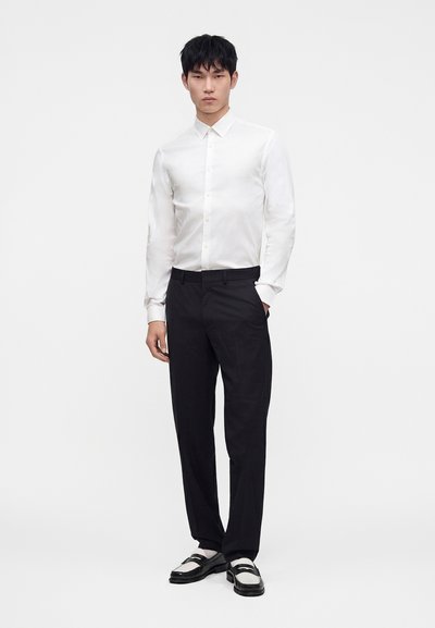 Man wearing a white button-up shirt, black trousers, and black and white loafers standing against a plain light background.