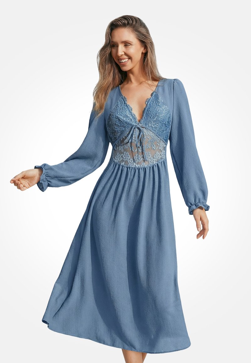 Donna sorridente che indossa un vestito midi a maniche lunghe blu con dettagli in pizzo e un laccio allo scollo a V, in piedi davanti a uno sfondo bianco neutro.