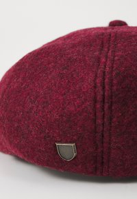 Red wool hat with a textured surface, featuring a metal shield logo and visible stitching detail on the crown.