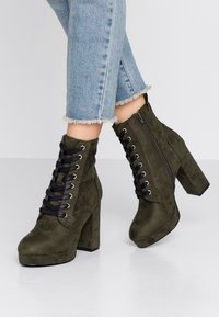Bottines en daim vert olive avec un talon épais, devant lacé, fermeture éclair sur le côté et accents en dentelle noire ; surface texturée et semelle à plateforme.