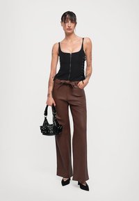 Schwarzes ärmelloses Oberteil mit Reißverschluss vorne und seitlichen Schnürungen, braune weite Hose, kleine schwarze Tasche mit Nieten und schwarze spitze Absätze.
