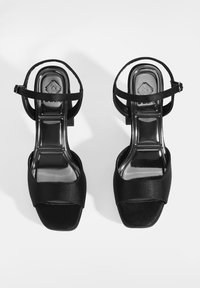Schwarze Satin-High-Heelsandalen mit offener Zehe, breitem Vorderriemen und schmalem Knöchelriemen mit Schnalle, die auf einer reflektierenden Oberfläche ruhen.