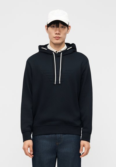 Giovane uomo che indossa una felpa nera con coulisse bianche, jeans scuri e un cappello bianco, su uno sfondo chiaro e uniforme.