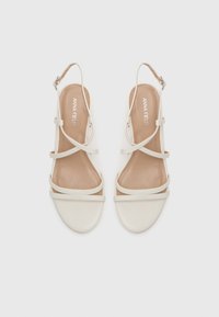White slingback sandals with crossed straps, a rounded toe, and a smooth texture. Soft beige insole. Buckle closure at the heel.